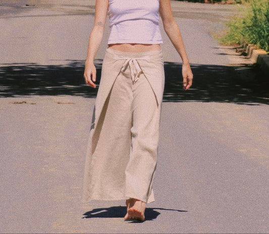 A woman walking on a sunlit paved path surrounded by trees, wearing a light lavender sleeveless crop top and loose beige wide-leg pants.