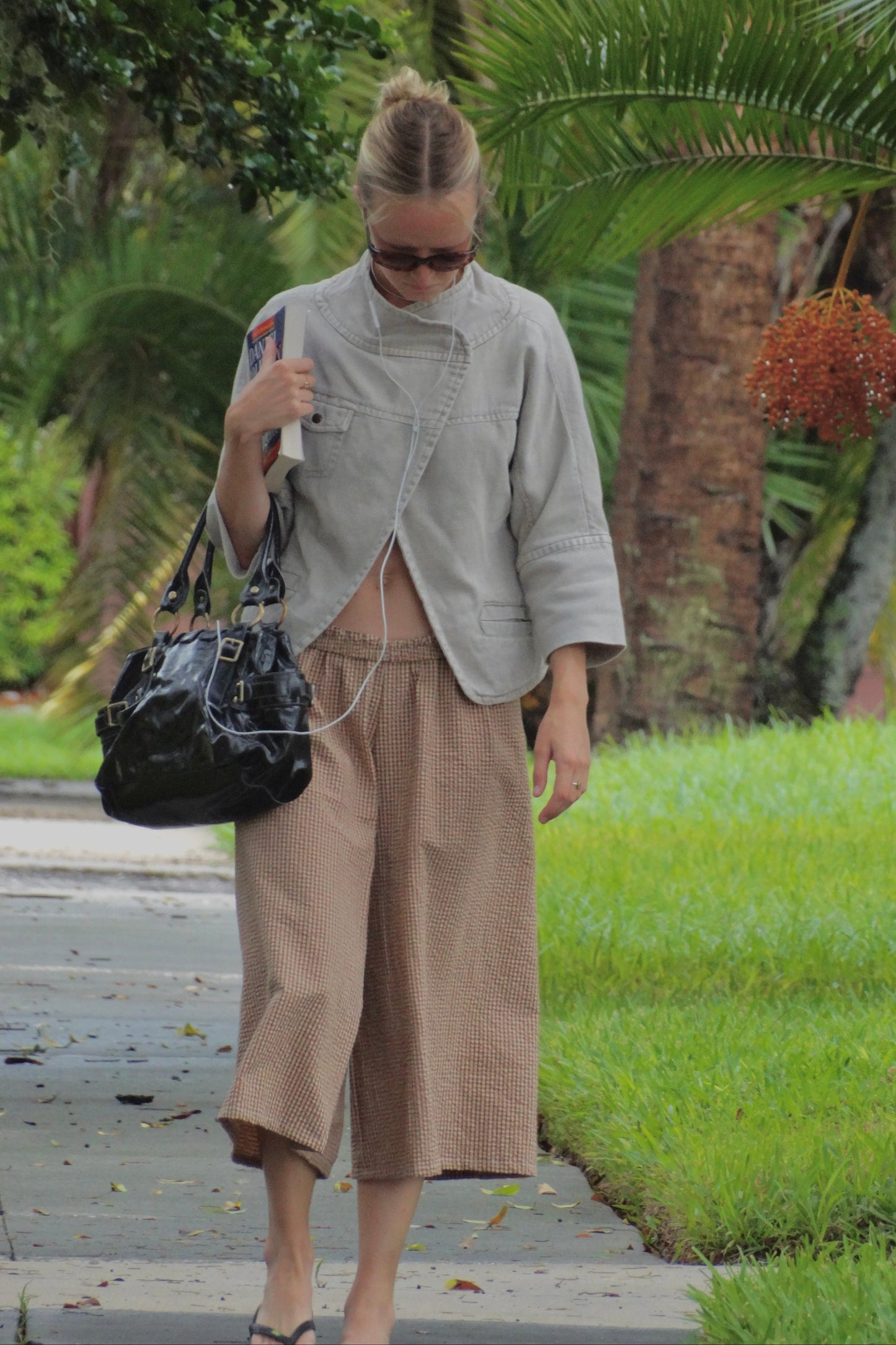 A woman walking outdoors on a paved path wearing a light gray cropped jacket, beige wide-leg cropped pants, and carrying a black shoulder bag.
