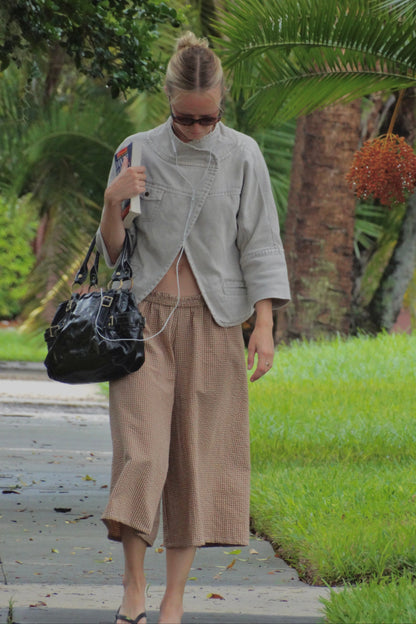 A woman walking outdoors on a paved path wearing a light gray cropped jacket, beige wide-leg cropped pants, and carrying a black shoulder bag.