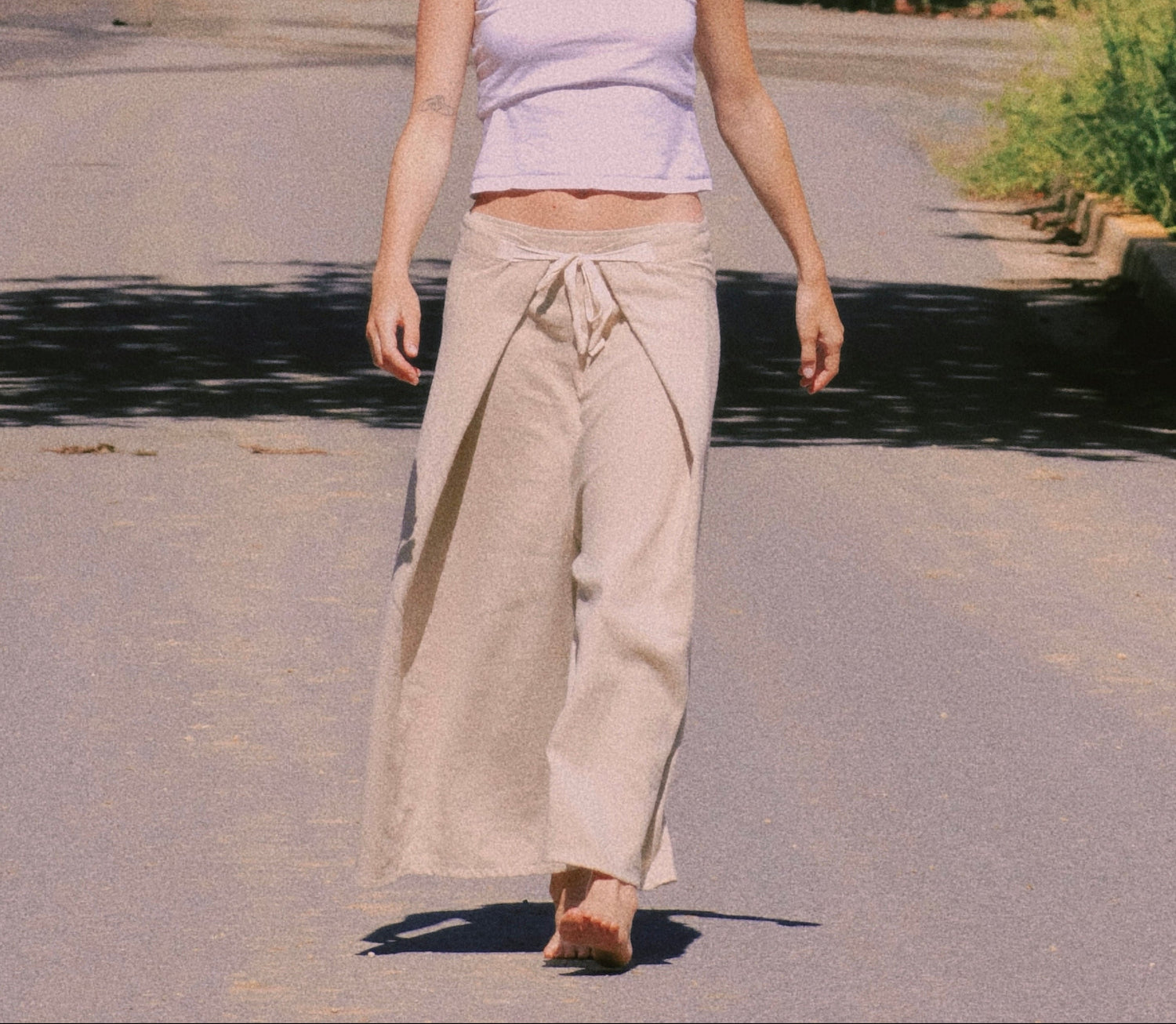 A woman walking on a sunlit paved path surrounded by trees, wearing a light lavender sleeveless crop top and loose beige wide-leg pants.